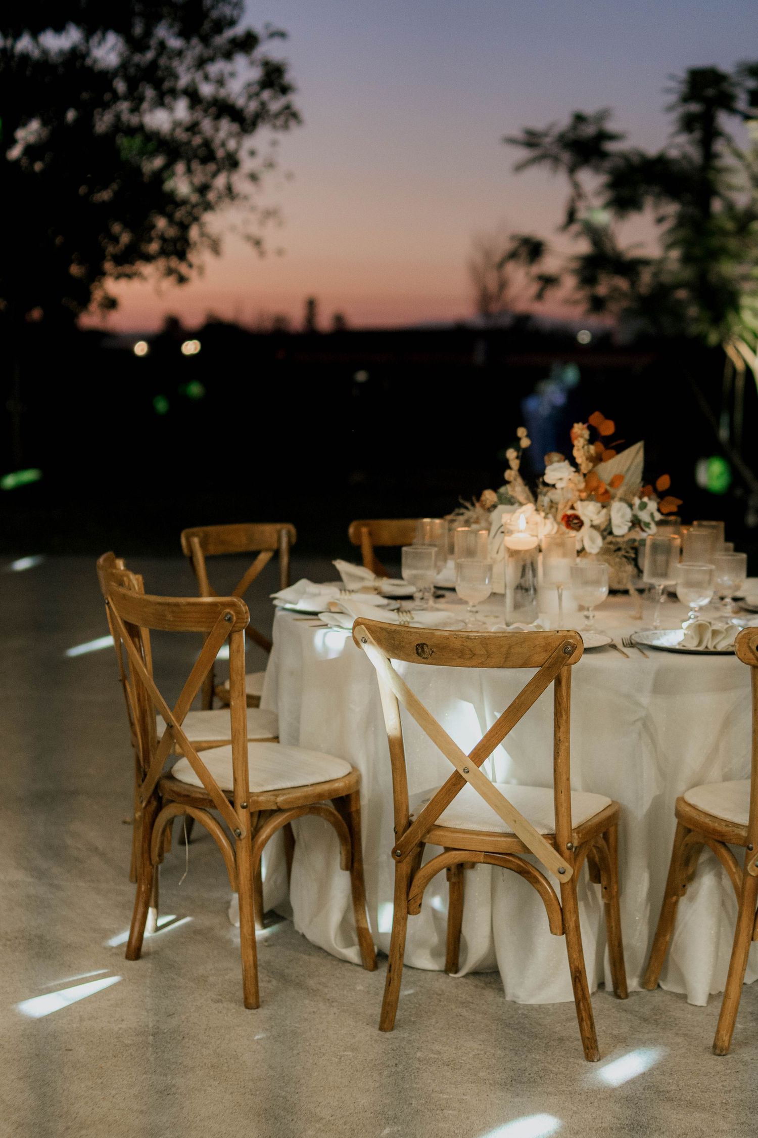 Elegant outdoor wedding reception setup with round tables, white linens and wooden cross-back chairs at sunset.