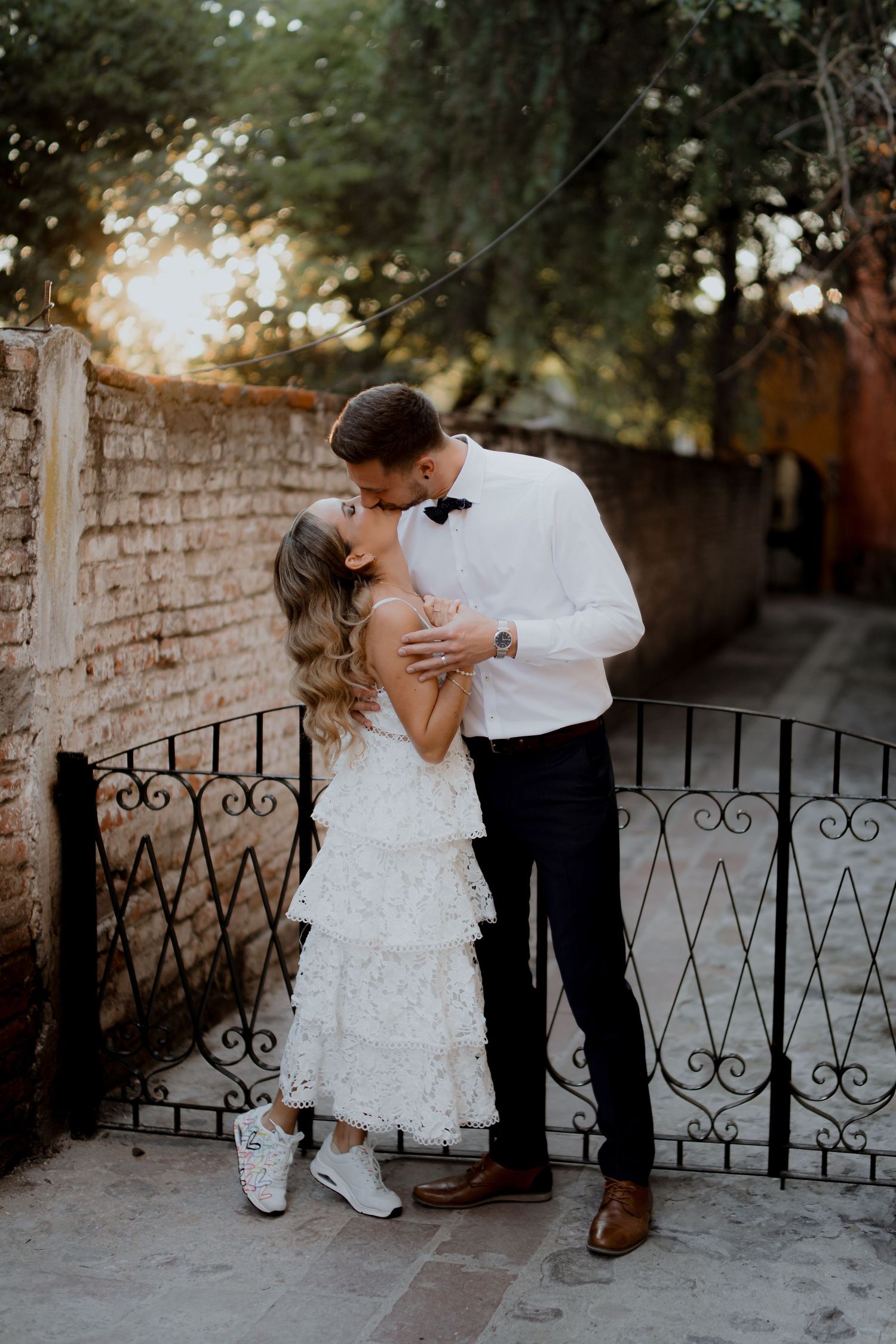 A romantic moment between a couple in formal attire by a rustic brick wall and iron gate.