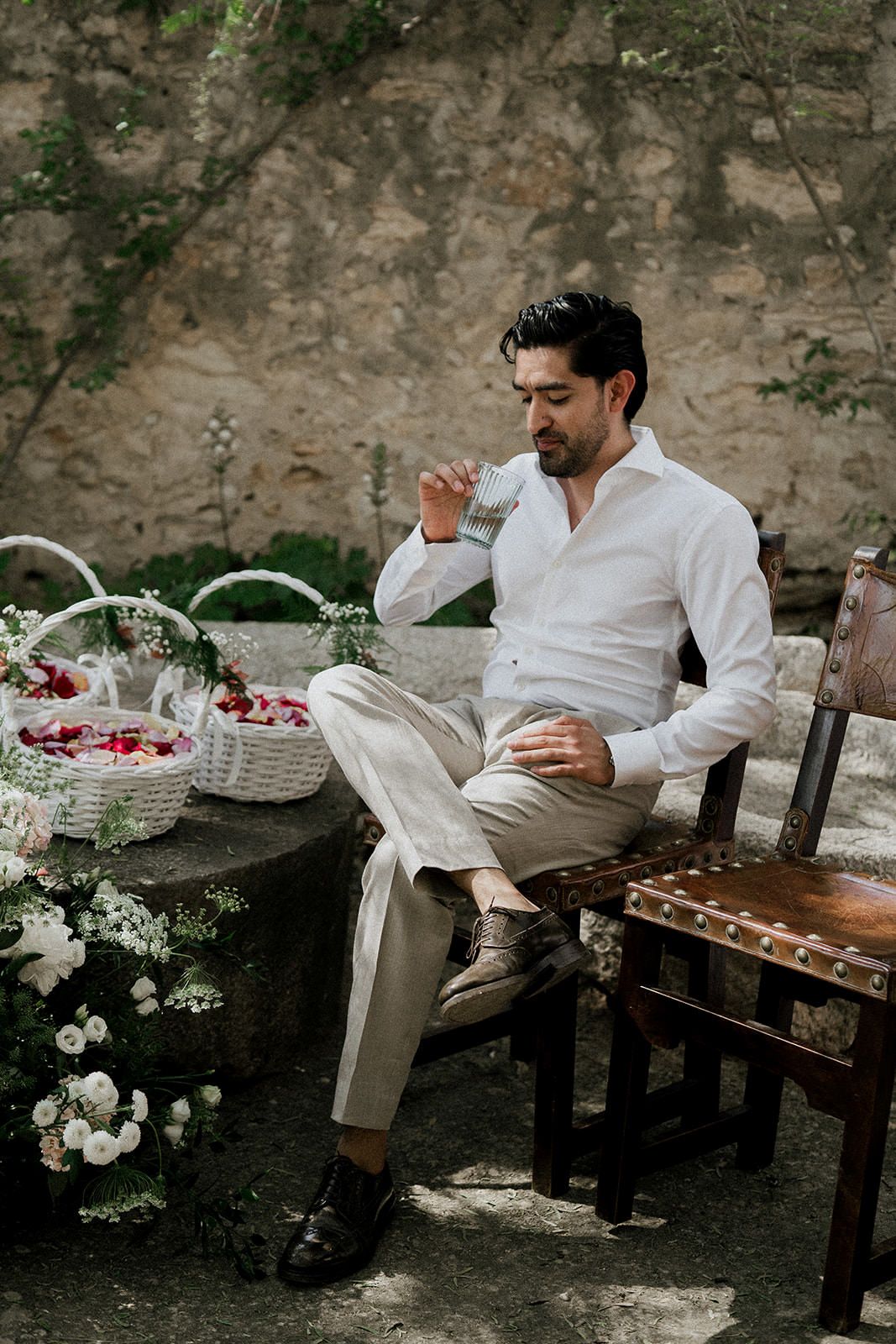 A person in white clothing sits on a wooden bench in a garden setting with flower baskets and sunlit stone walls.