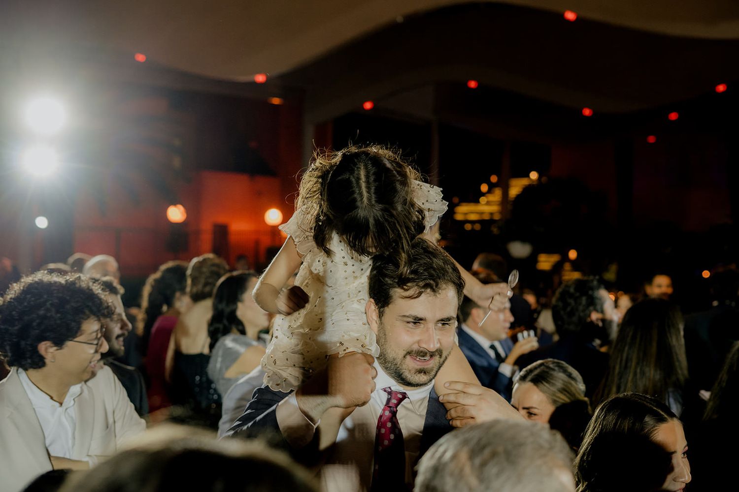 Excited flower girl sits on groomsman shoulders celebrating joyfully on crowded wedding reception dance floor.