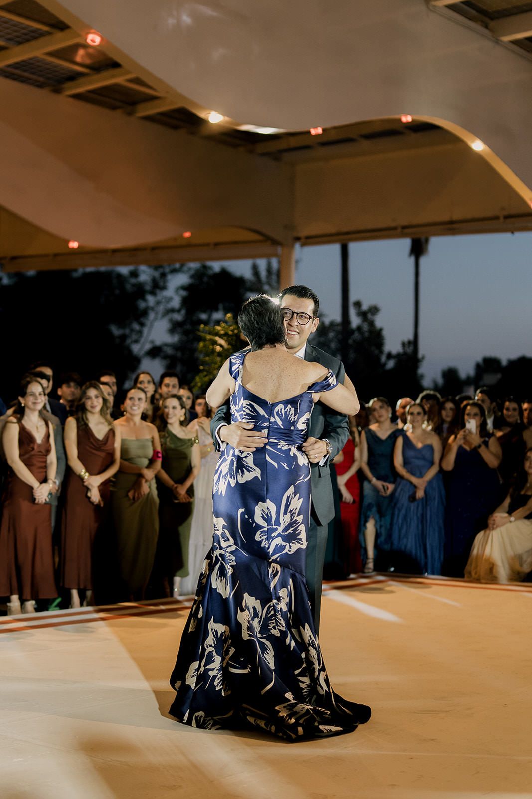 Groom embracing mother in blue floral mermaid gown during emotional mother-son dance at outdoor wedding reception.