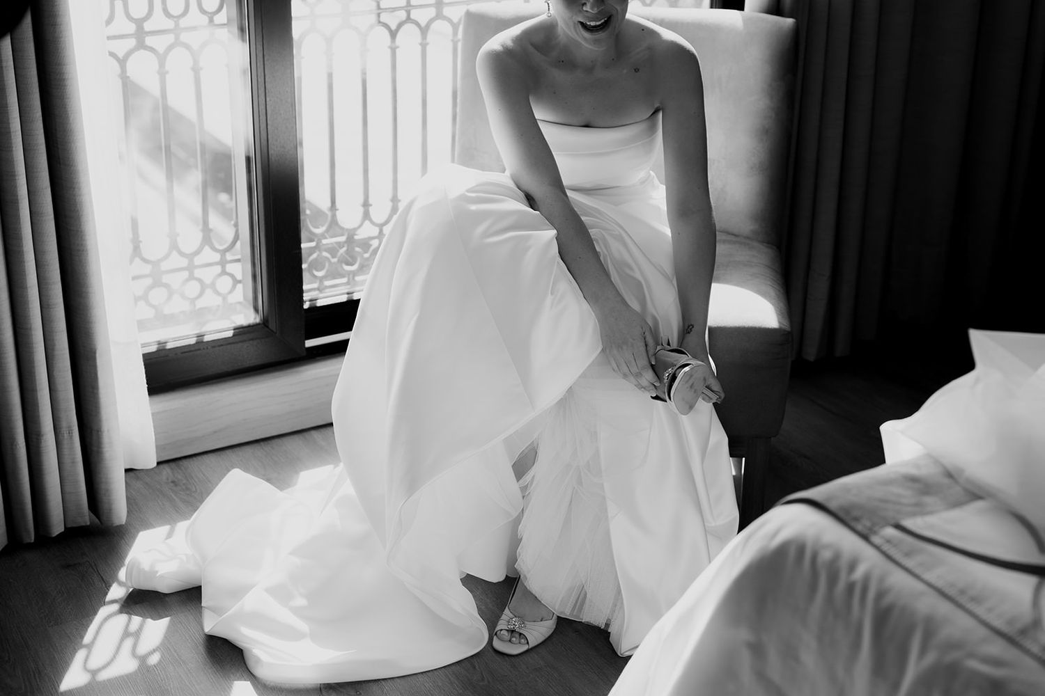 Bride in a strapless white wedding gown adjusting her heels in a sunlit room, black and white photo.