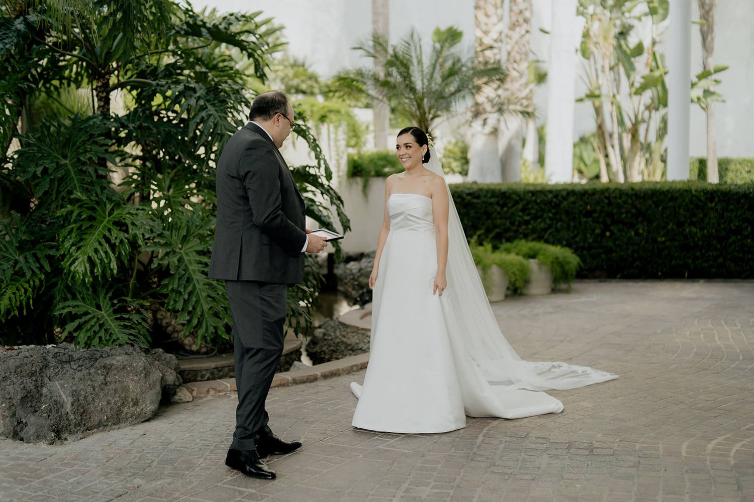 Bride in white strapless gown and groom in dark suit share a first look moment in an elegant outdoor courtyard.