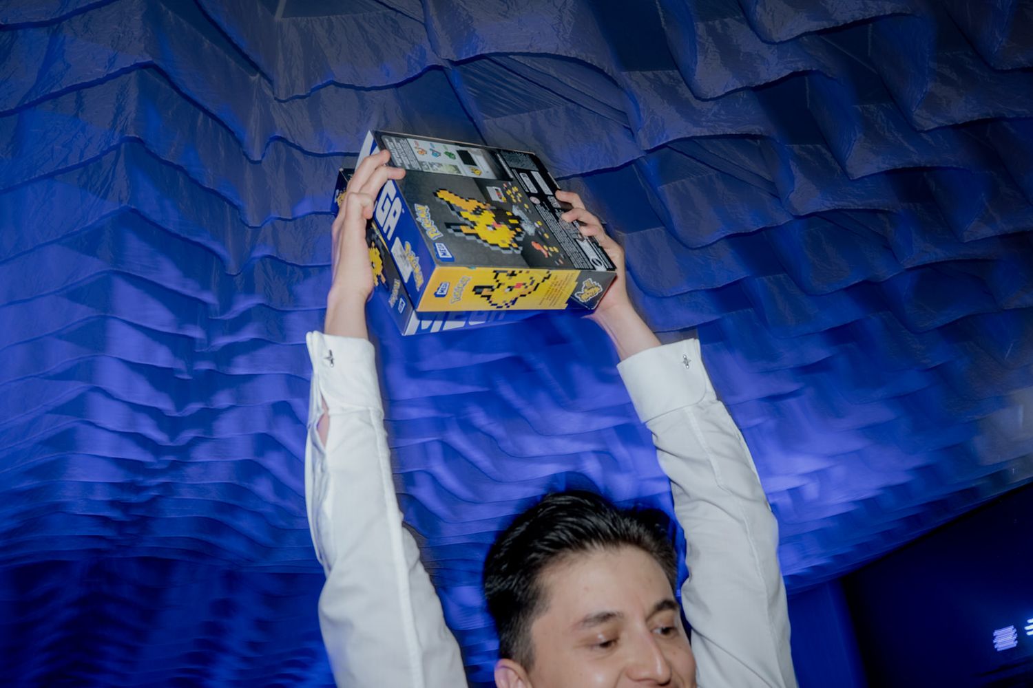 Man in white shirt raises a colorful box overhead at a blue-lit wedding celebration.