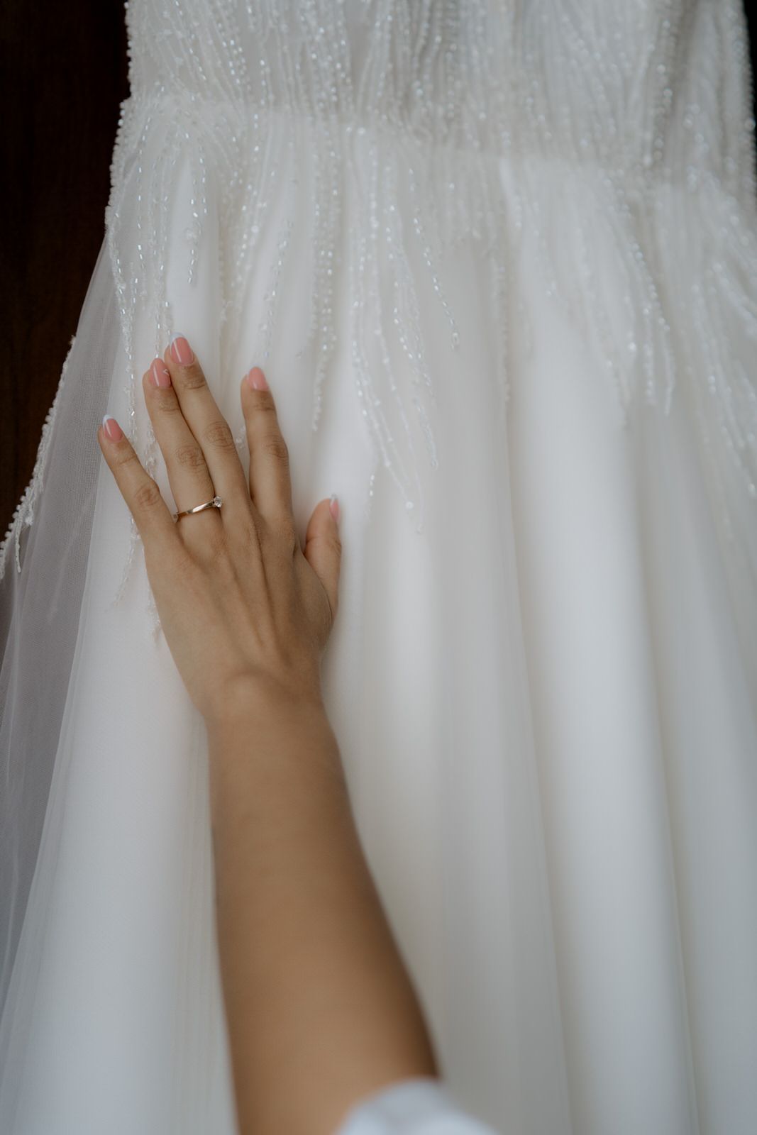 Bride's hand with gold wedding ring touching beaded white wedding dress.