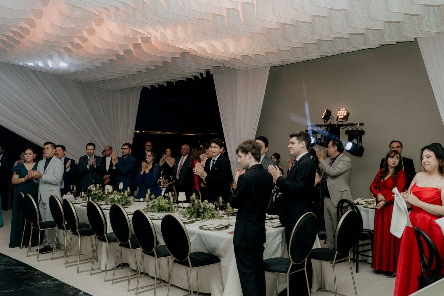 Elegantly dressed wedding guests applaud at reception with long decorated dining tables.