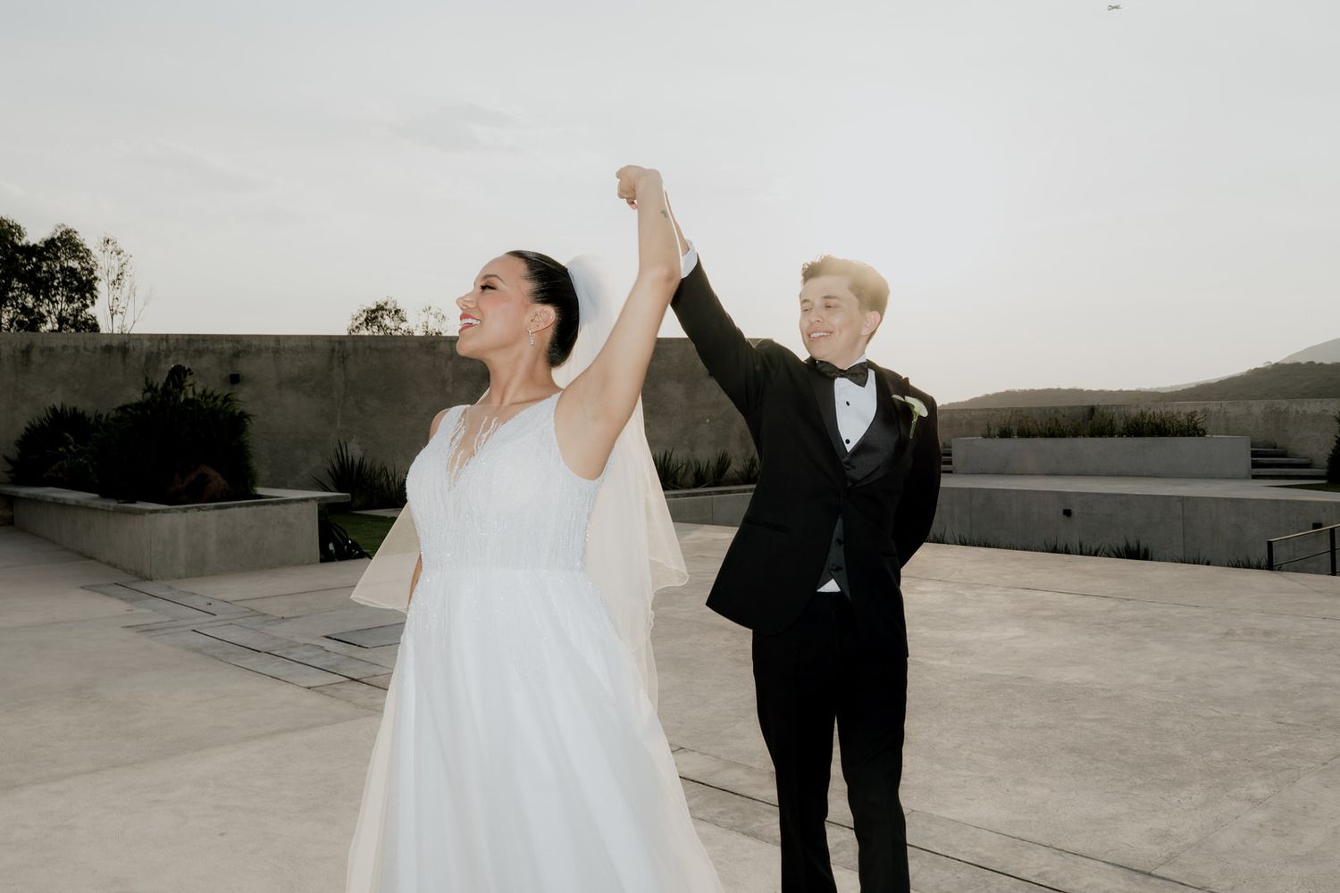 Joyful bride and groom celebrate on outdoor terrace, groom raising brides arm in triumph against twilight sky.