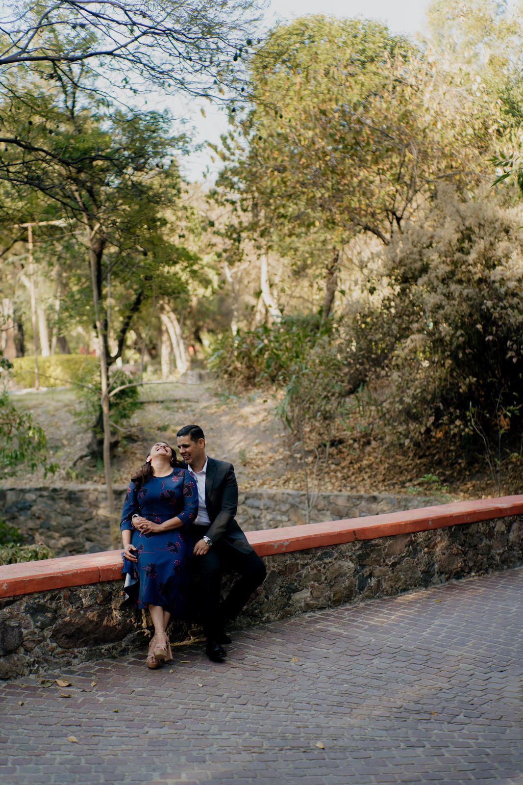 Couple seated on a brick wall surrounded by lush trees and greenery.