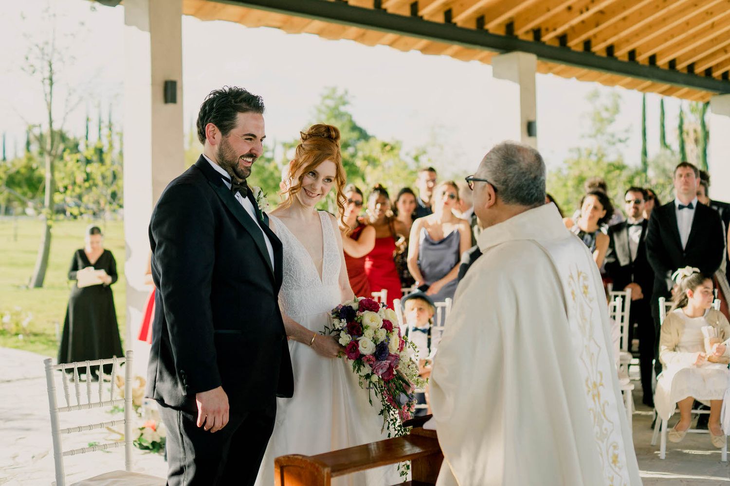 Outdoor wedding ceremony under wooden pavilion with guests witnessing special moment.
