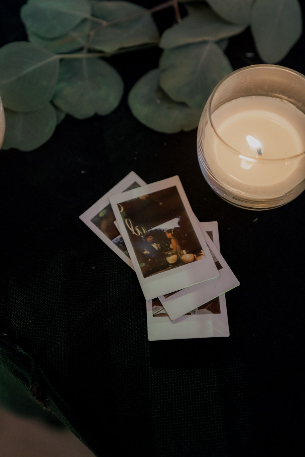Polaroid photos lay next to a white candle and eucalyptus leaves on a dark surface.