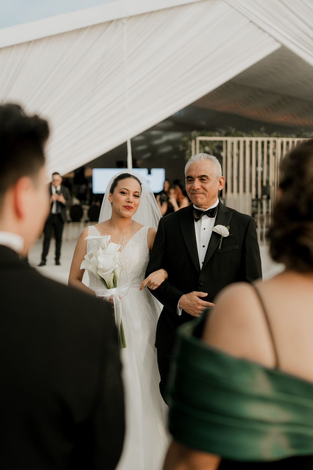 Bride in white gown holding white bouquet walks down aisle with older man in tuxedo at outdoor ceremony.