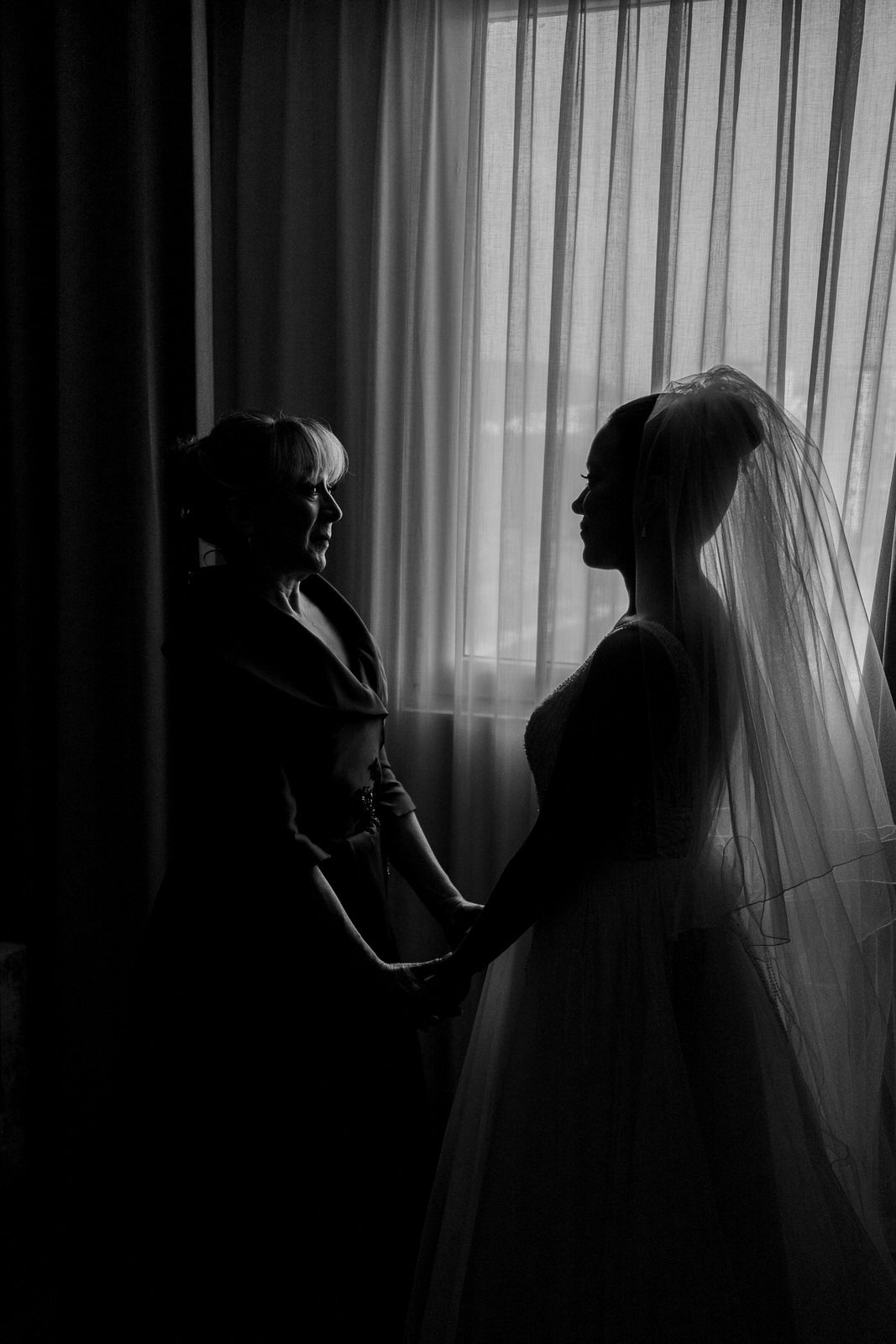 Black and white silhouette of bride in veil and wedding gown holding hands with woman near curtained window.