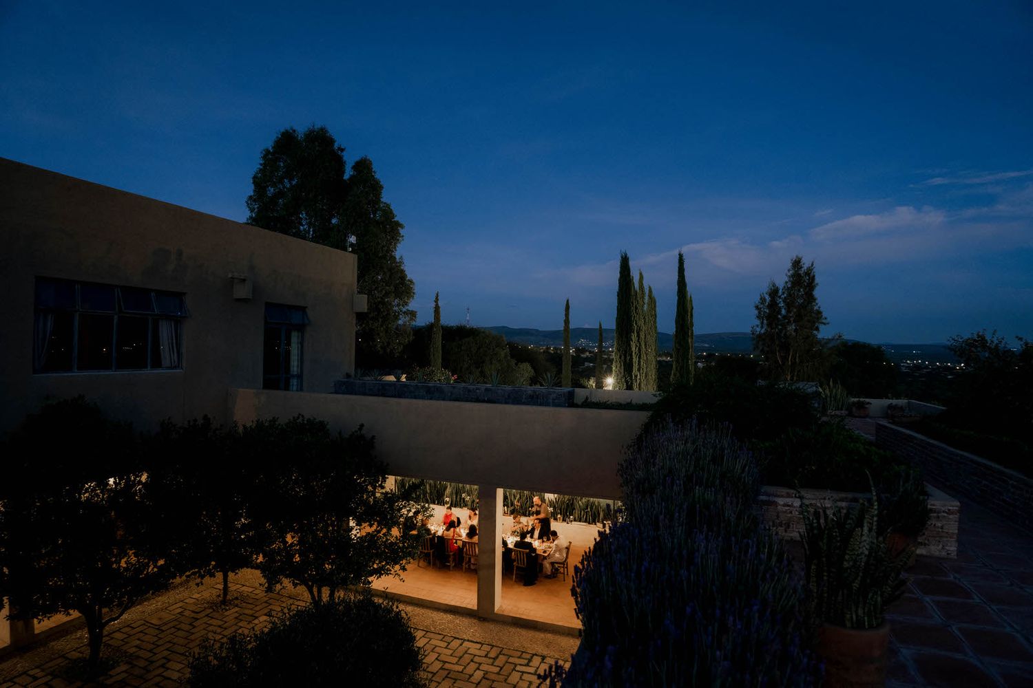 Evening outdoor reception venue with warm lighting and cypress trees against blue twilight sky.