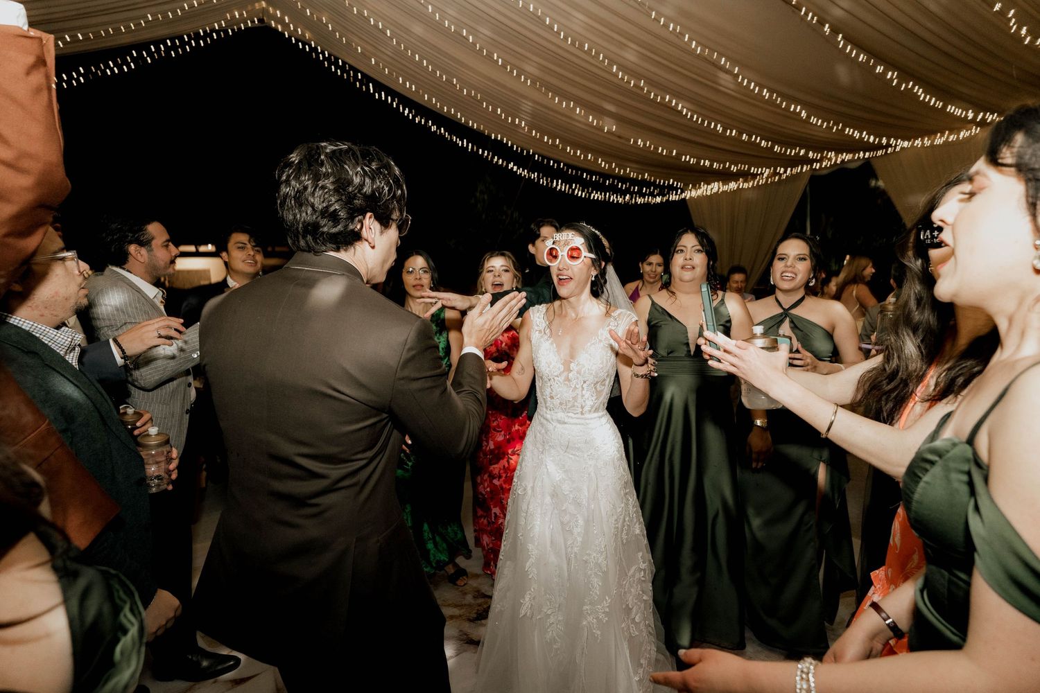 Wedding reception guests dancing in circle under decorative ceiling lights.