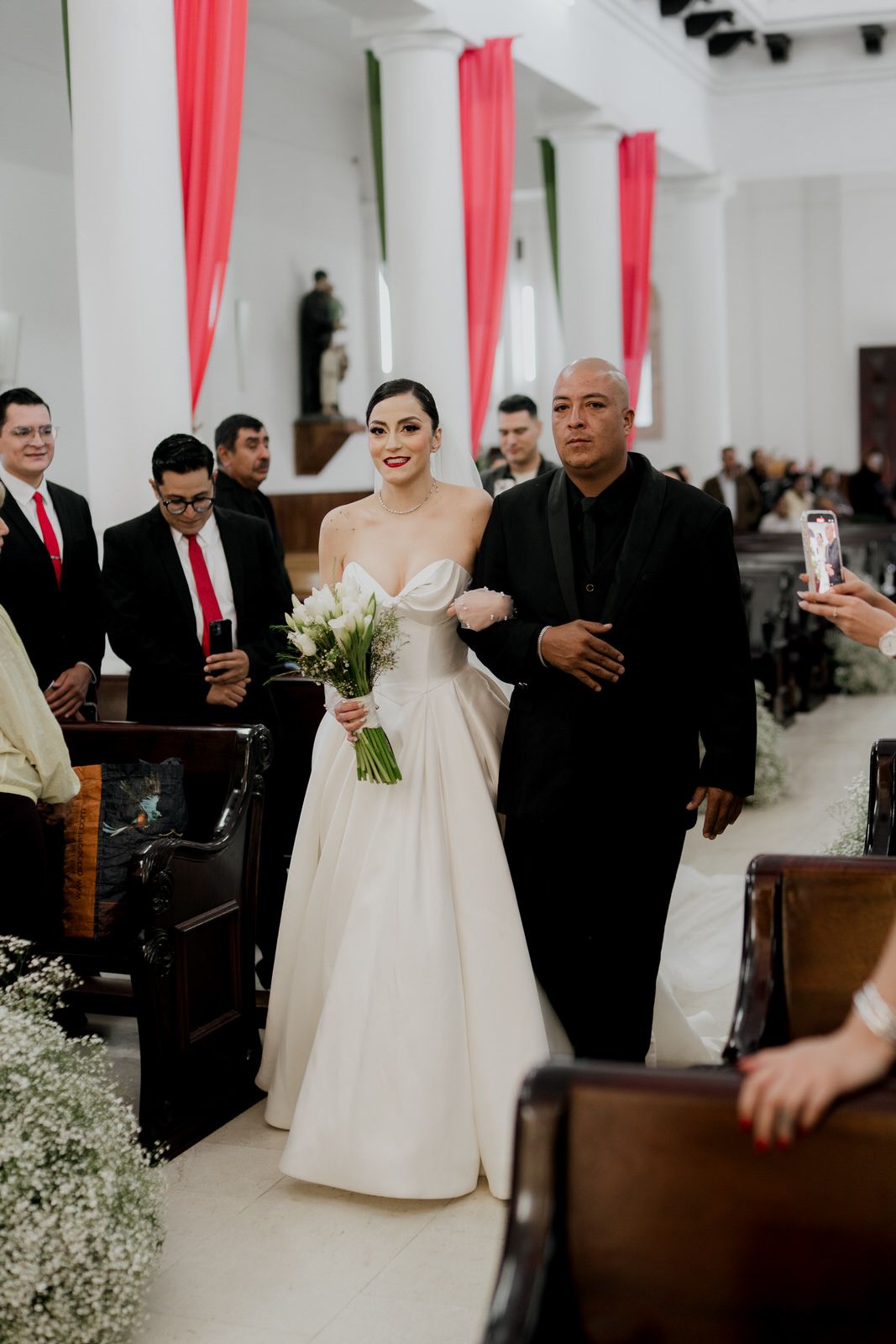 Wedding party walking down church aisle decorated with white flowers and red accents.