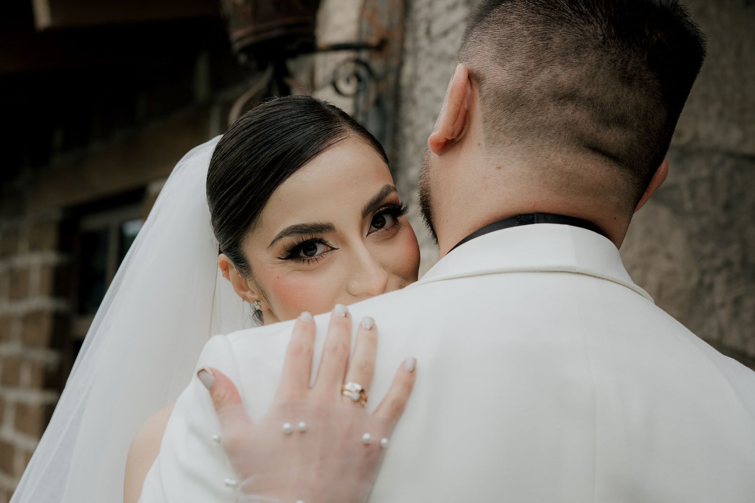 Artistic portrait of bride with veil showing engagement ring against white suit.