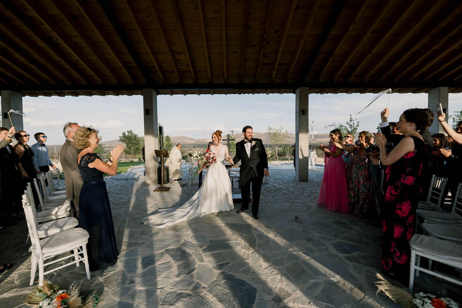 A wedding ceremony procession in a sunlit outdoor pavilion with guests watching from both sides.