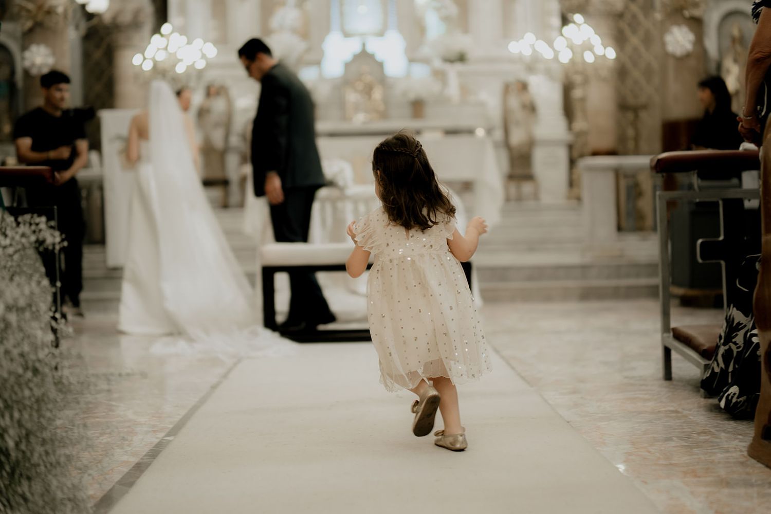 A flower girl in a white lace dress walks down the aisle toward the altar at a church wedding ceremony.