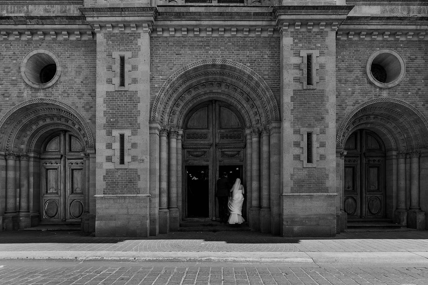 Black and white photo of a bride in a white gown standing at the grand entrance of a historic stone church.