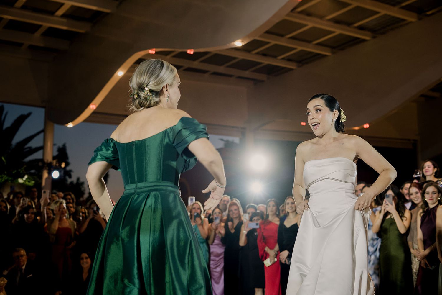 Two women in elegant gowns, one green and one white, face each other at a formal event with a crowd in the background.