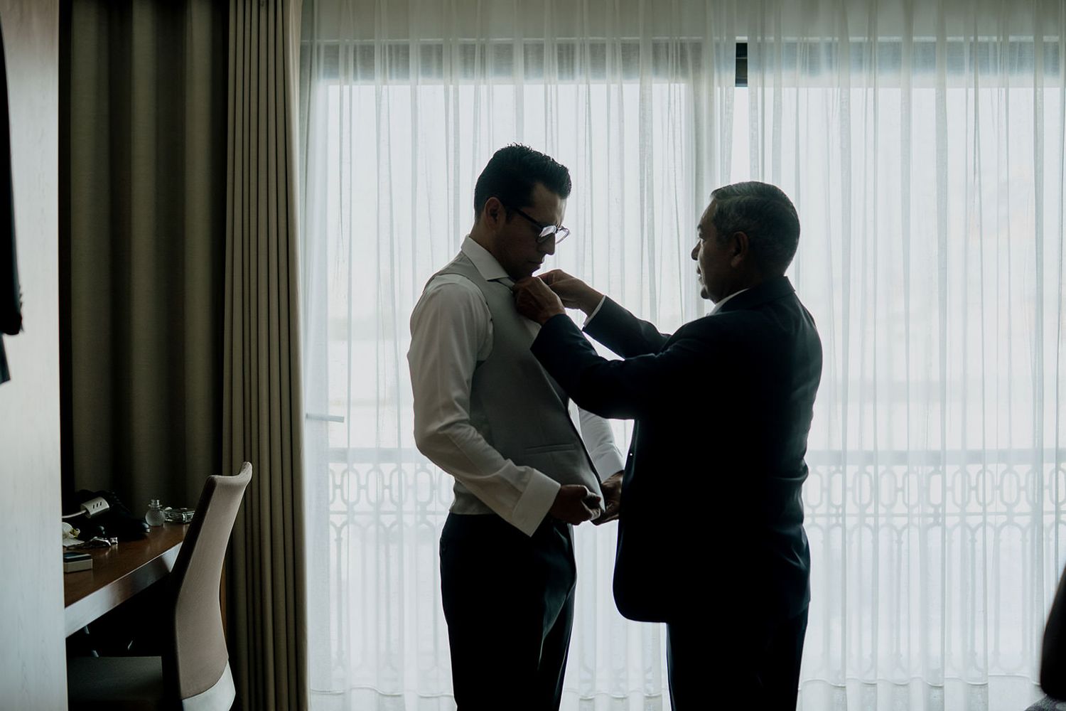 Two men in silhouette, one helping the other adjust his tie near a bright window.