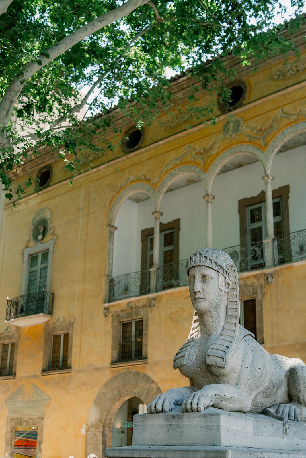 Stone sphinx statues line a yellow building facade with windows and trees overhead in an ornate architectural setting.