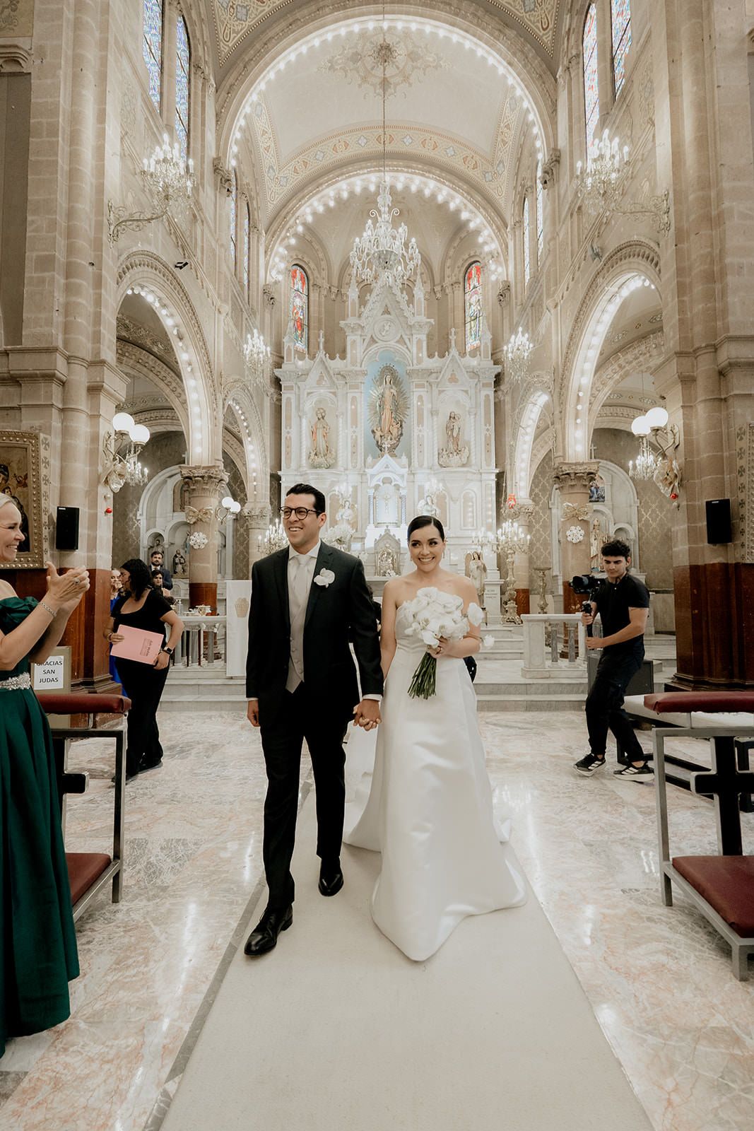 Bride and groom walk down the aisle of an ornate church with chandelier and white altar.