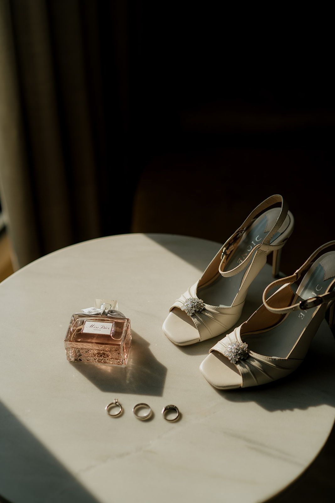 Bridal flat lay with white heels, wedding rings, and Dior perfume on a marble table in soft natural light.