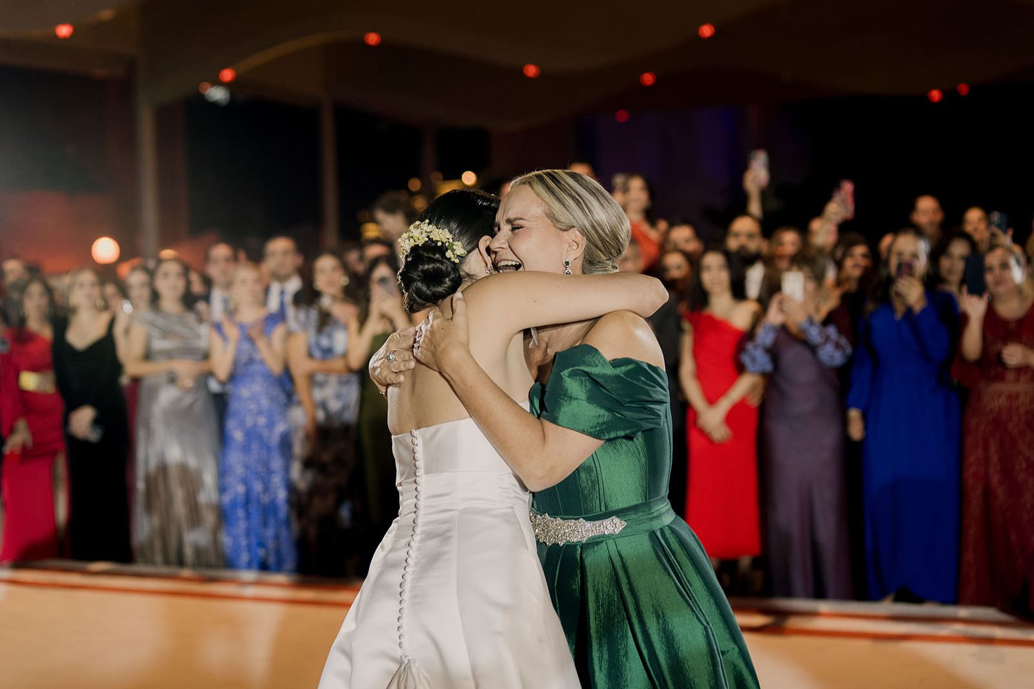 Bride and woman in green dress share emotional hug on wedding dance floor with guests applauding in background.