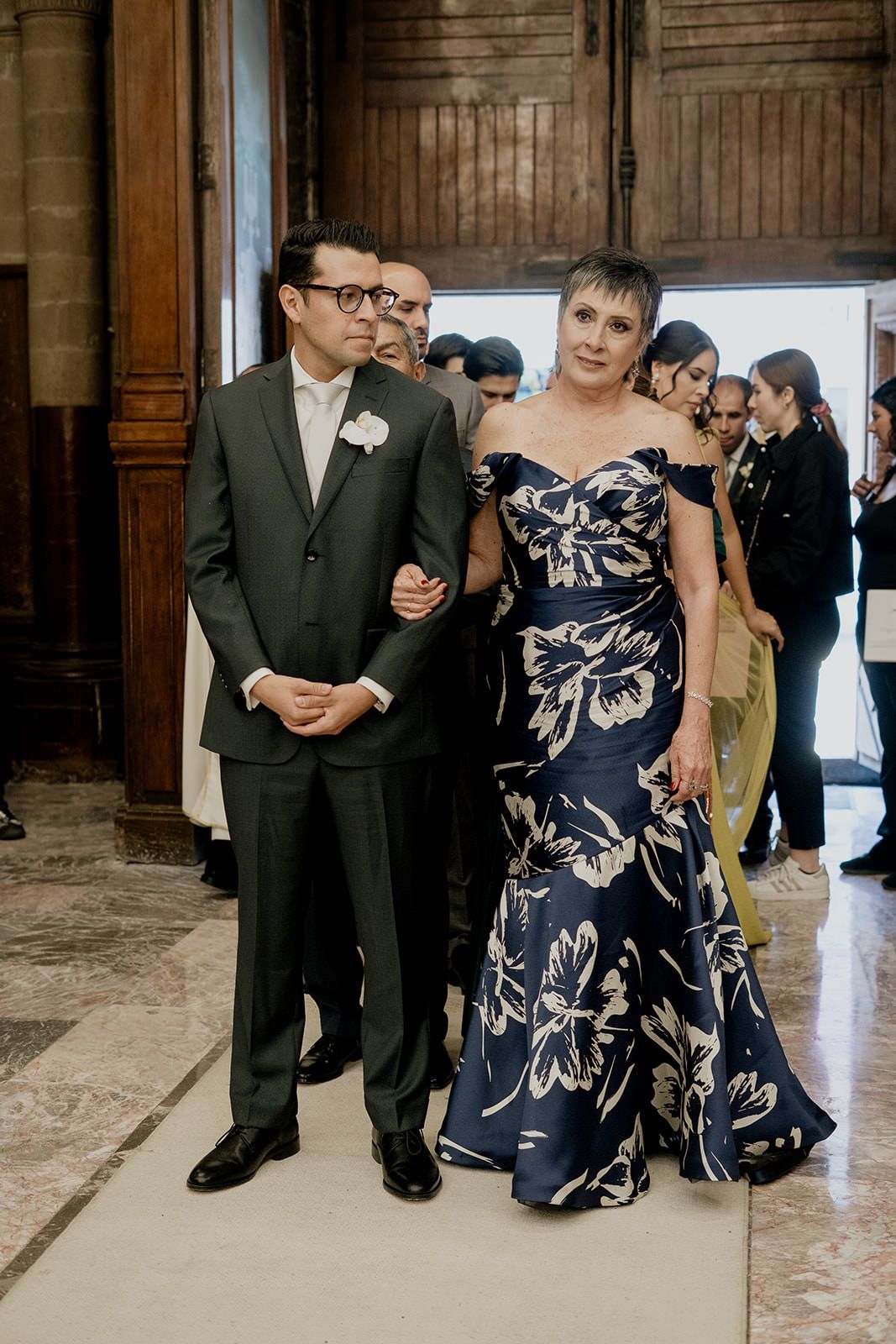 Groom in dark suit escorts elegantly dressed woman in navy floral gown down church aisle at wedding ceremony.