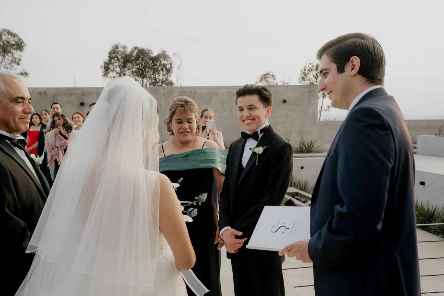 Bride and groom exchange vows at outdoor wedding ceremony with smiling guests and officiant holding white book.