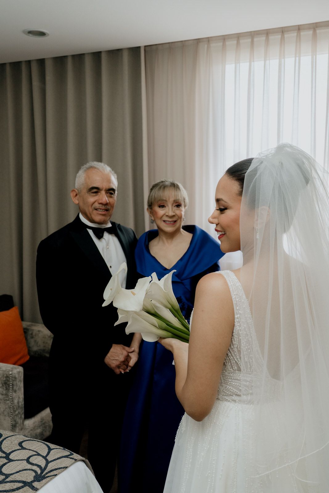 Bride in white gown holding calla lilies with parents in formal attire.