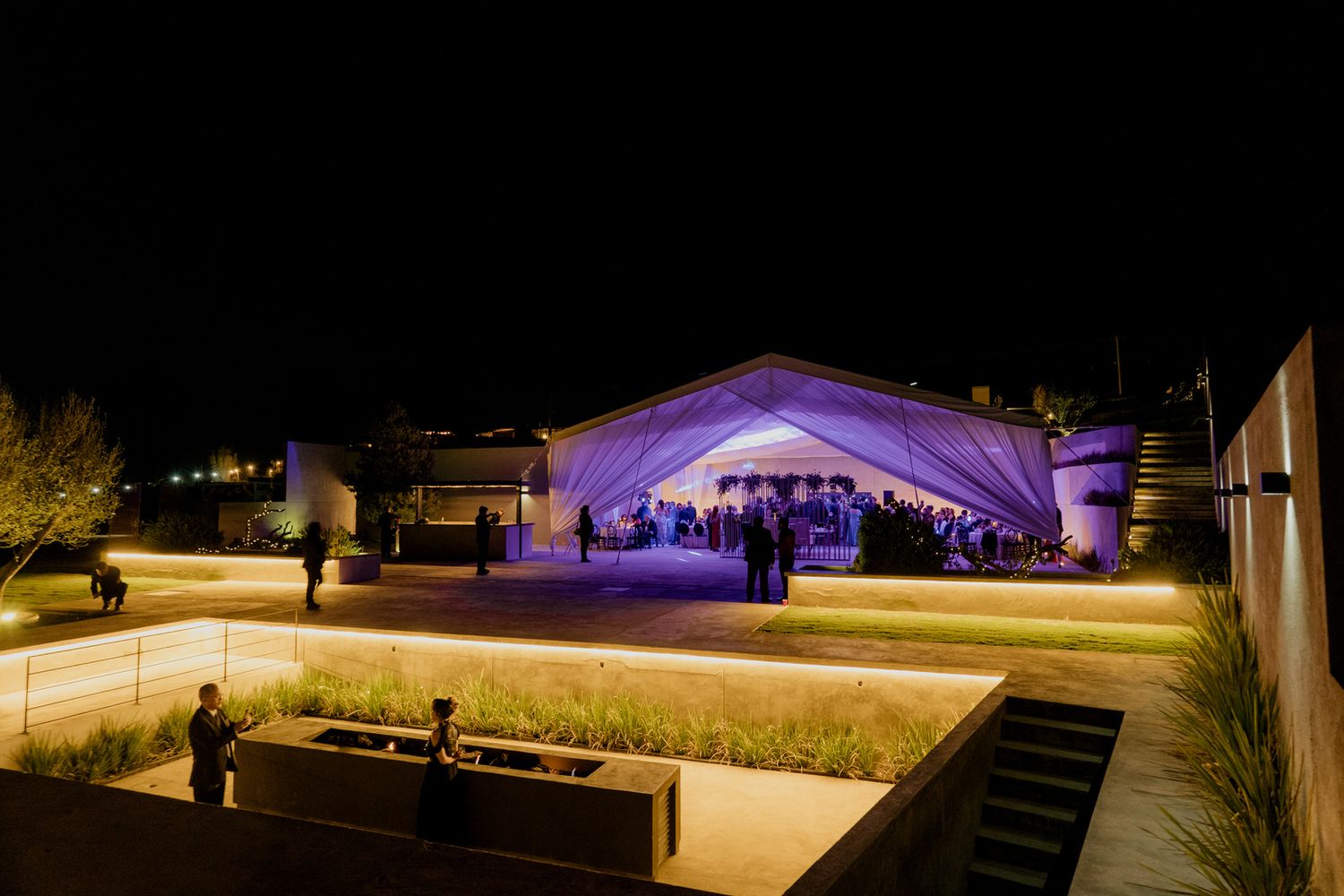 Elegant outdoor wedding venue illuminated at night with purple lighting and guests gathered under a large white tent.