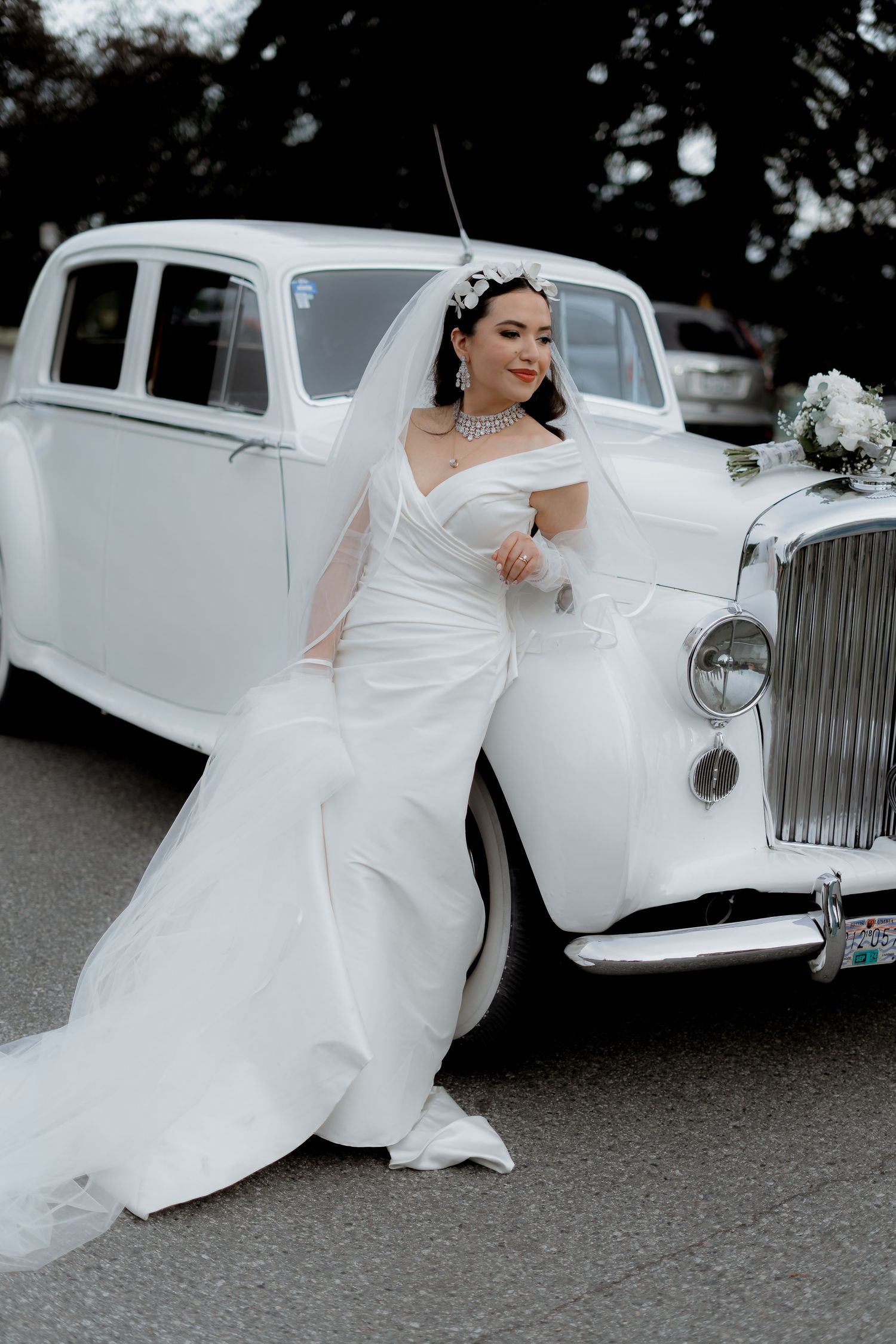 A bride in an elegant white gown poses beside a classic vintage white car.