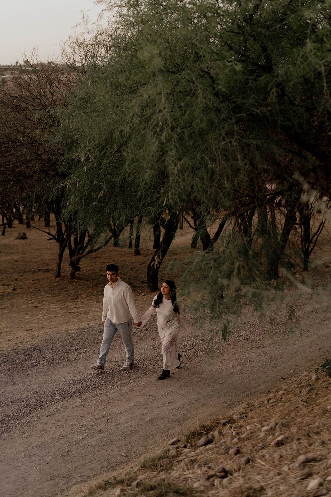 Two people walk together on a desert trail surrounded by trees.