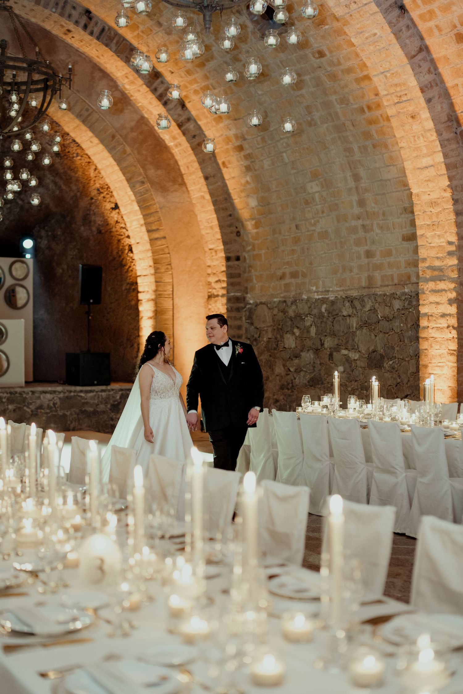 Wedding reception setup in a rustic brick vault with white table settings and hanging lights.