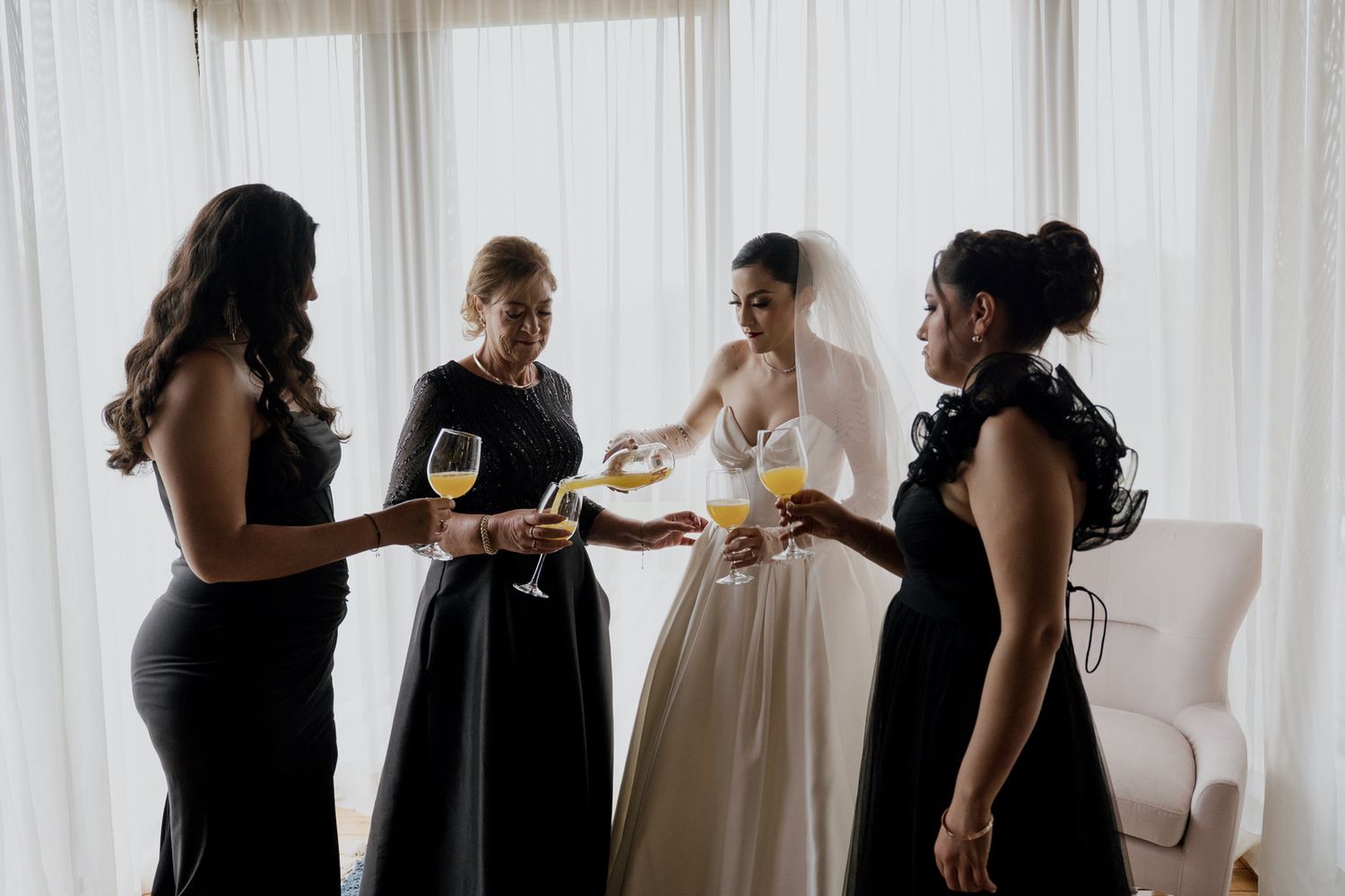 Wedding party in formal black dresses gather around person in white gown holding drinks.
