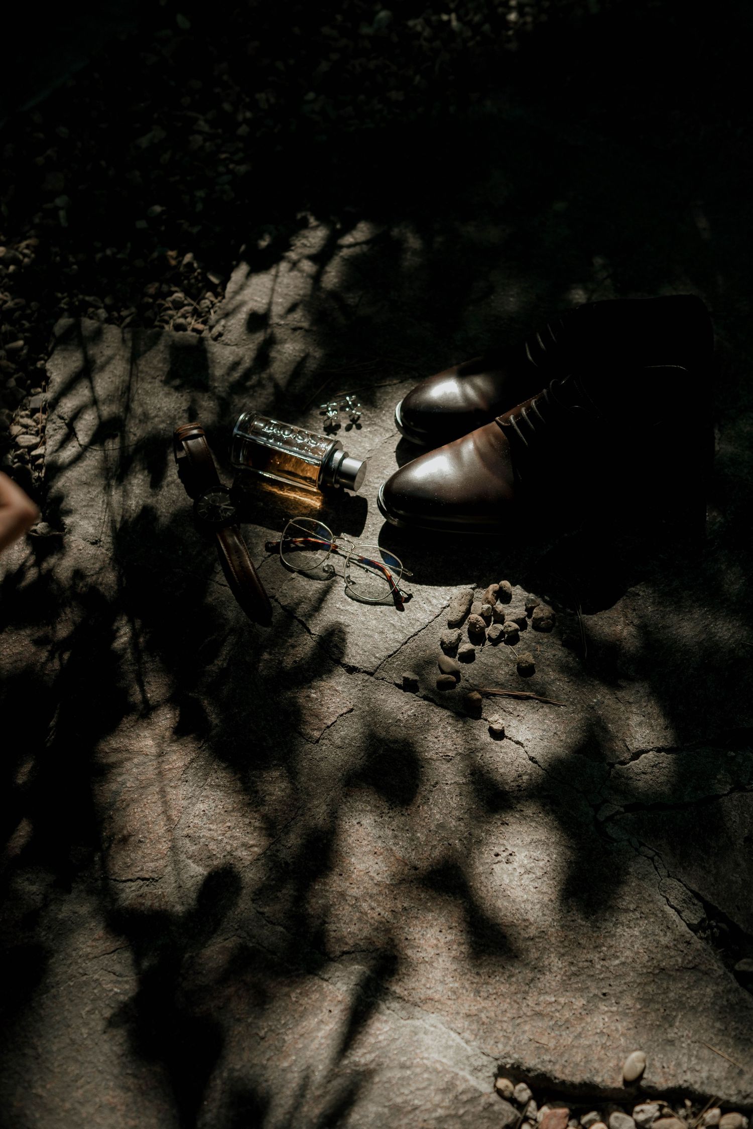 Elegant dress shoes and accessories arranged in dramatic lighting with dappled shadows.
