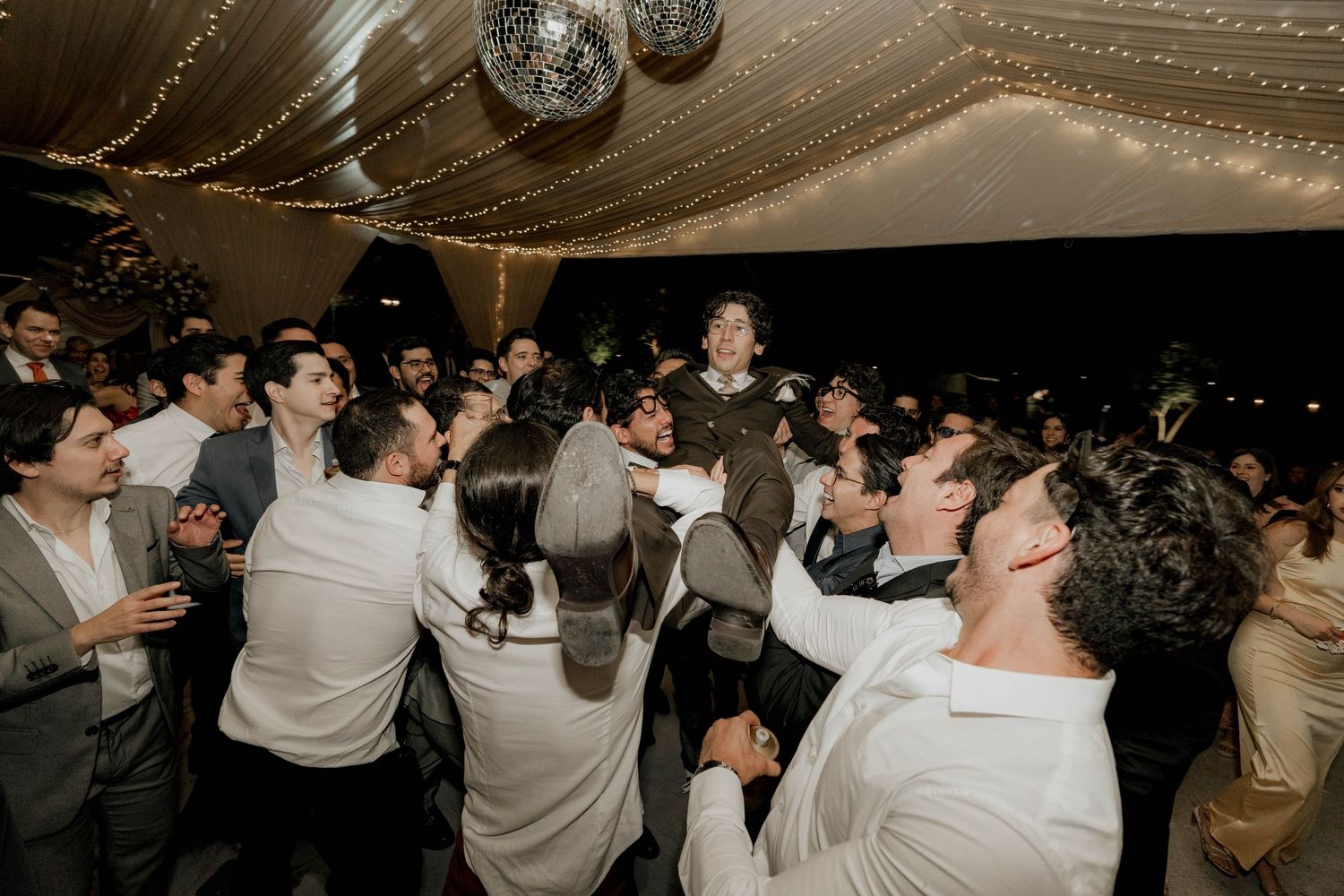 Crowd of wedding guests dancing enthusiastically under disco balls and fairy lights.