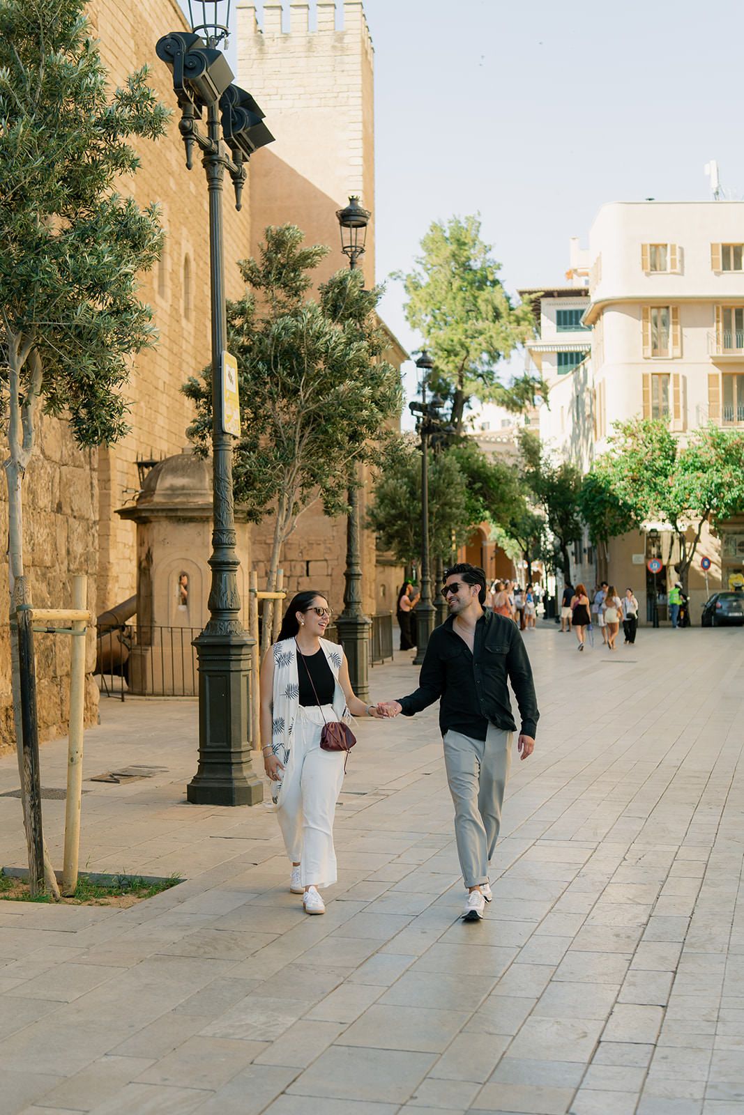 People walking along a stone-paved street with historic buildings and lamp posts in a Mediterranean setting.