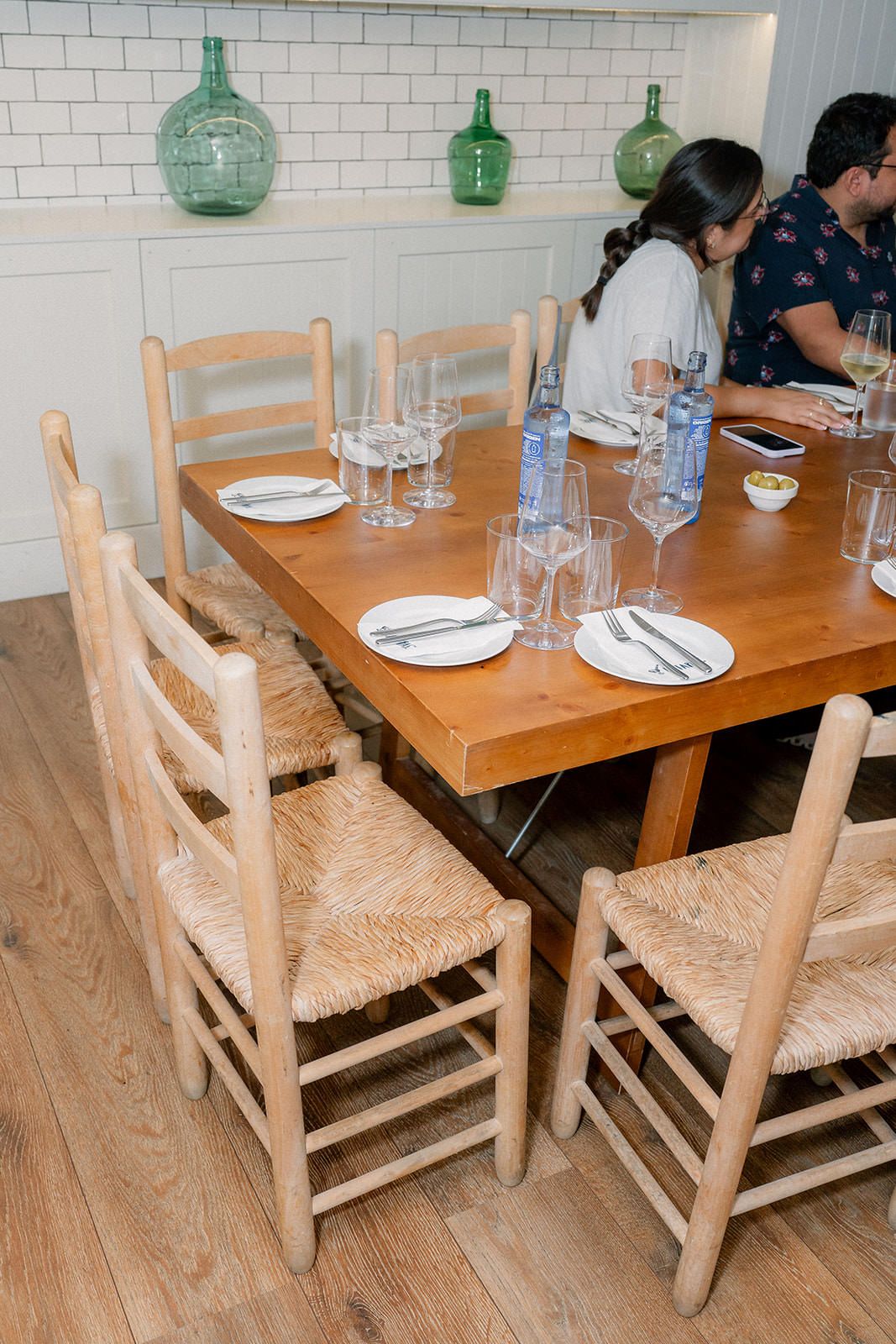 Rustic restaurant interior with wooden tables and woven straw chairs against white subway-tiled walls.