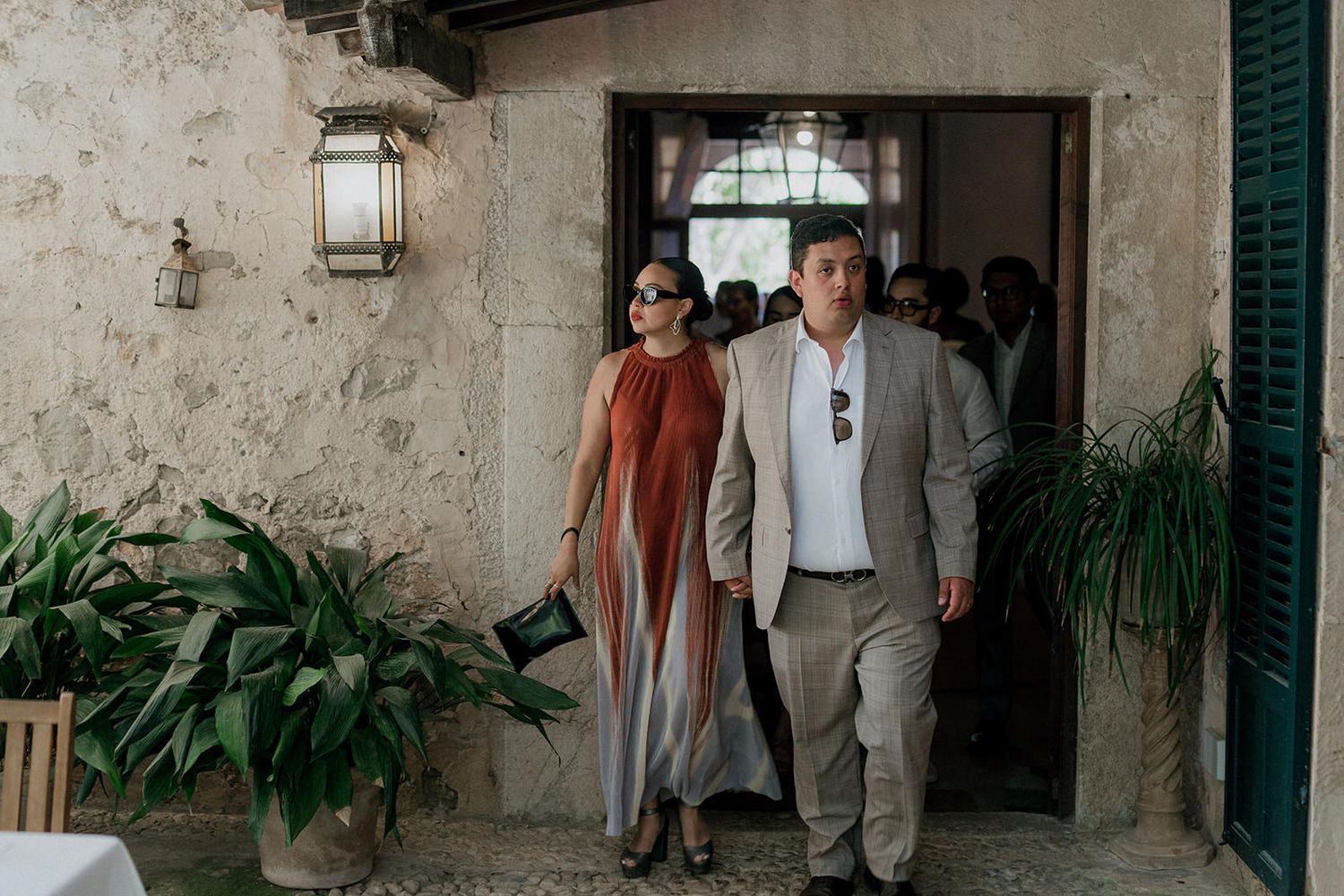 Wedding guests gather in a rustic stone courtyard with green shutters and potted plants.