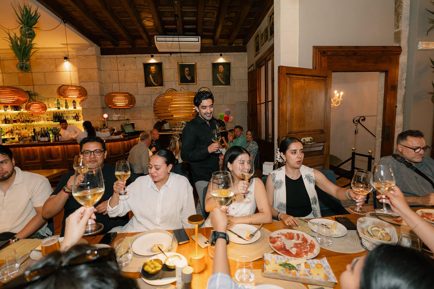 A lively dinner party scene with guests gathered around a wooden table in a warmly lit restaurant setting.