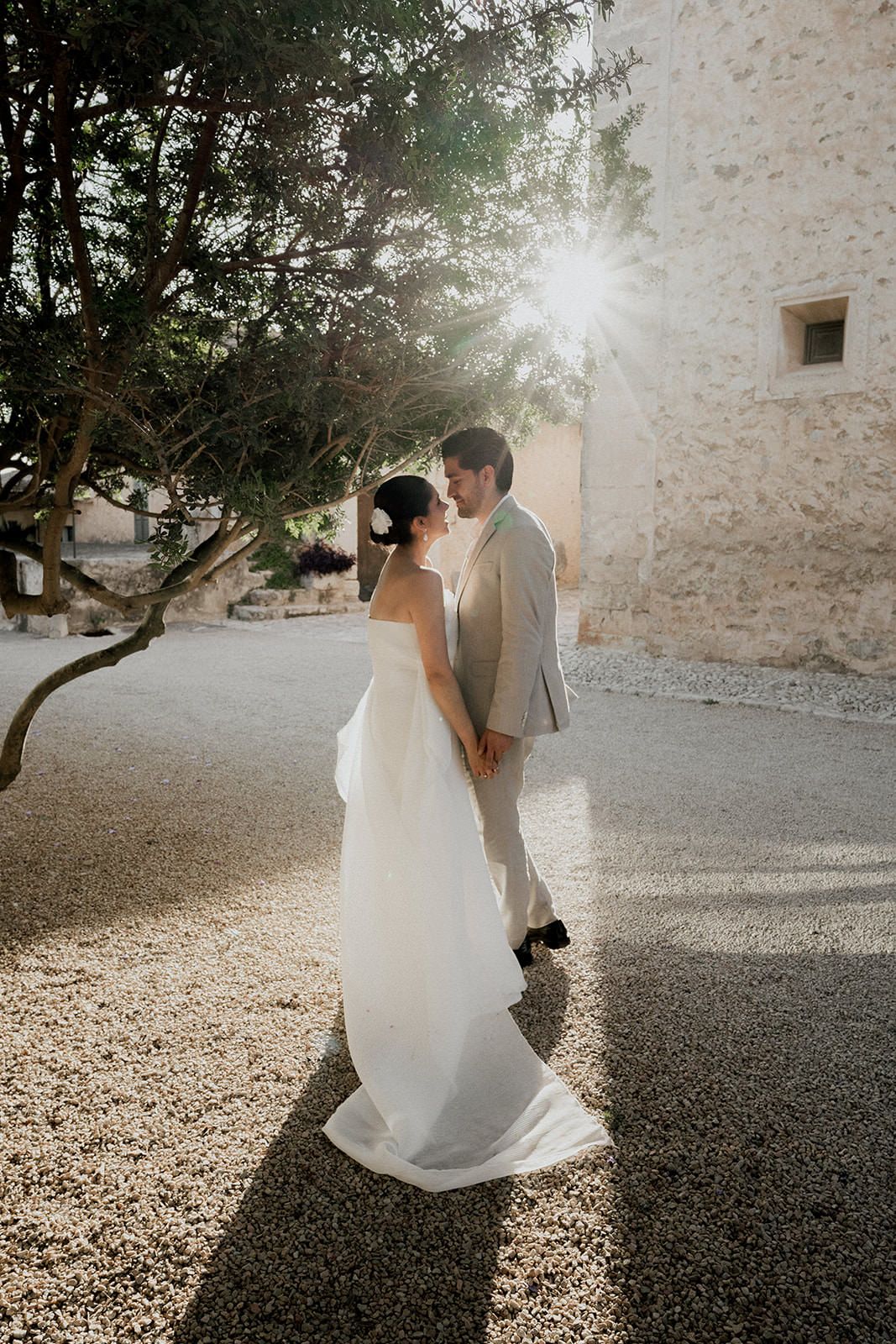 A couple in wedding attire dances together on a gravel courtyard beside a rustic stone wall in warm sunlight.