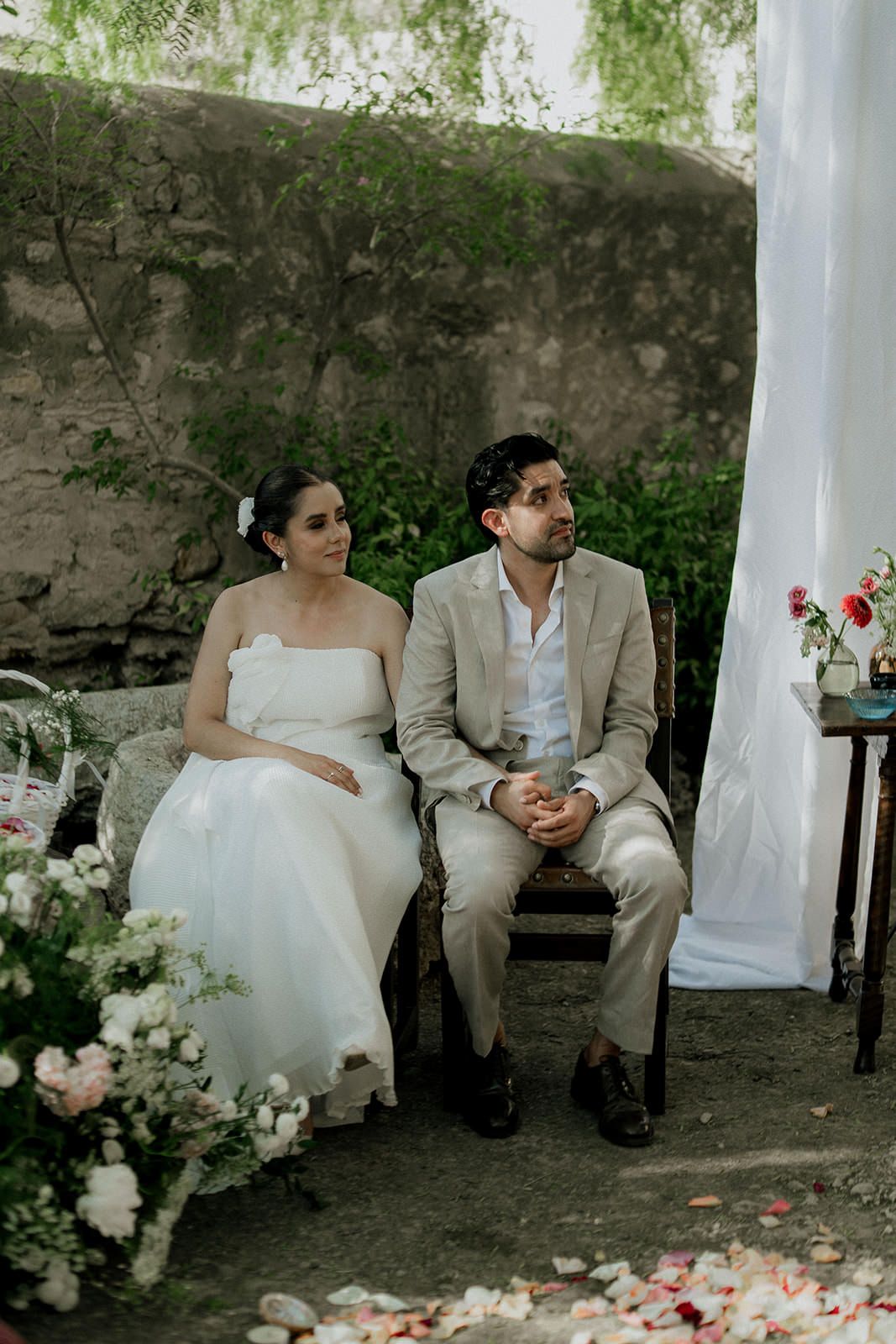A couple in wedding attire sits together on a bench under a white gazebo with rose petals scattered on the ground.