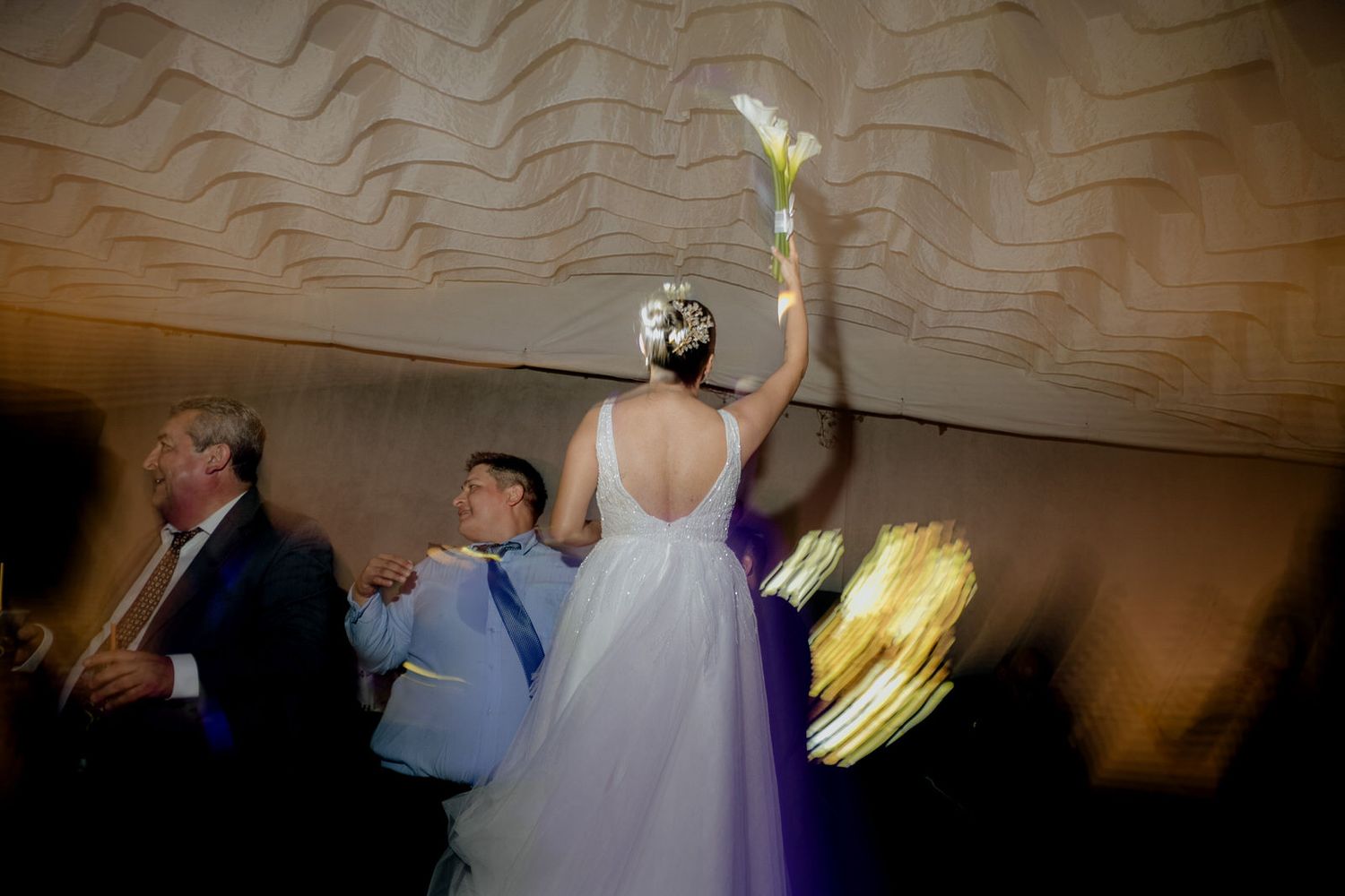 Bride in white backless gown raises bouquet on dance floor with guests at elegant wedding reception.