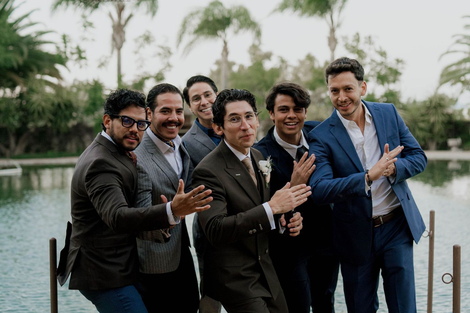 Groomsmen in suits share a candid moment of laughter by a waterfront palm tree setting.