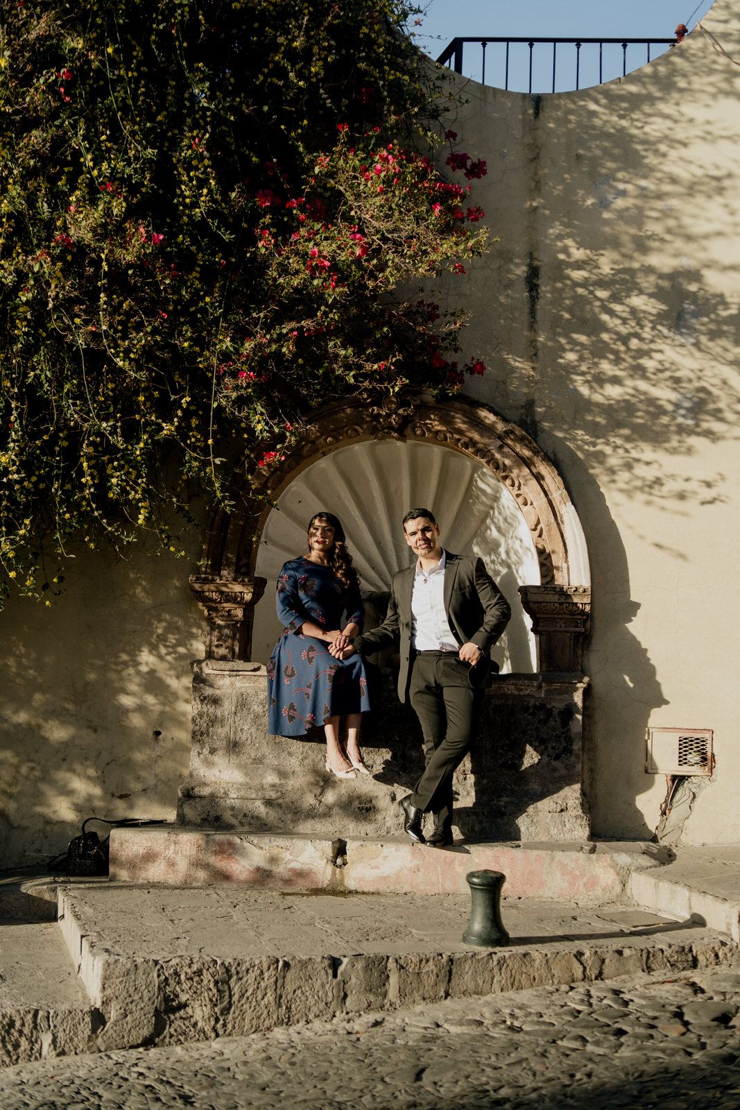 Couple stands beneath stone archway with flowering vines in colonial setting.