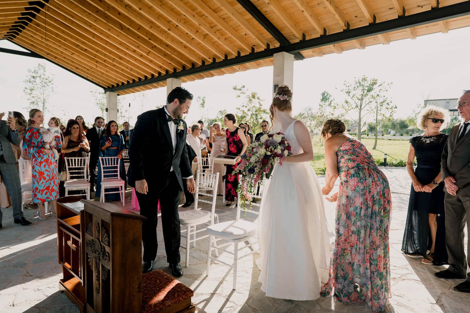 Wedding ceremony in progress under wooden pavilion with guests seated in white chairs.