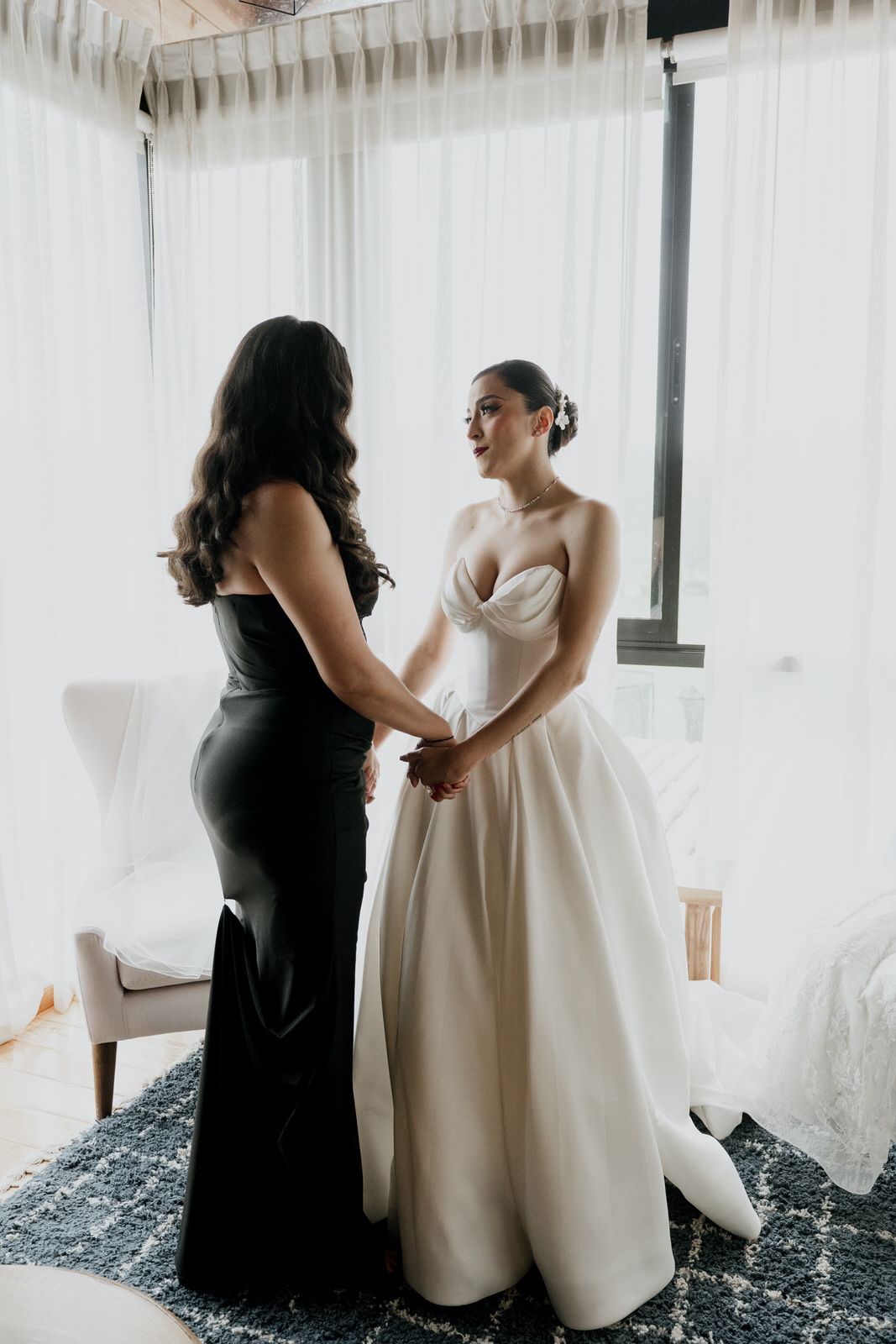 Two people share a moment during wedding preparations by white curtained windows.
